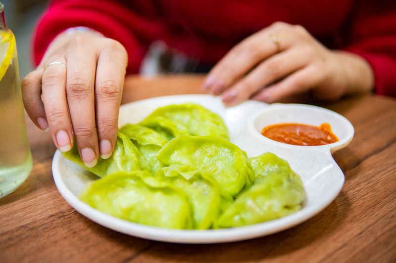 Close up indian young woman sitting in a restaurant and eating green color delicious Chinese steamed dumplings, dim sum, momo or jiaozi with red spicy chili sauce in white plate on wooden table and drinking Refreshing Mojito Cocktail.