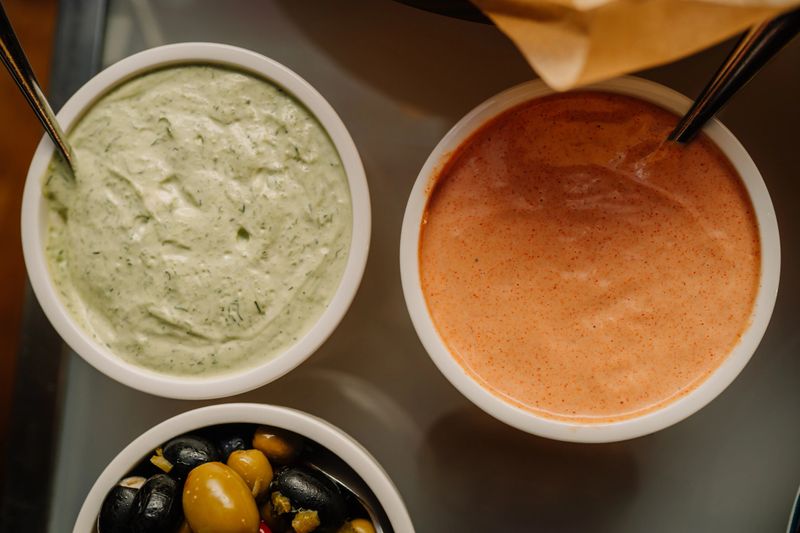 Two bowls of creamy saucesone green herb-based and one orange spicedserved with a side of mixed olives in a catering setup.