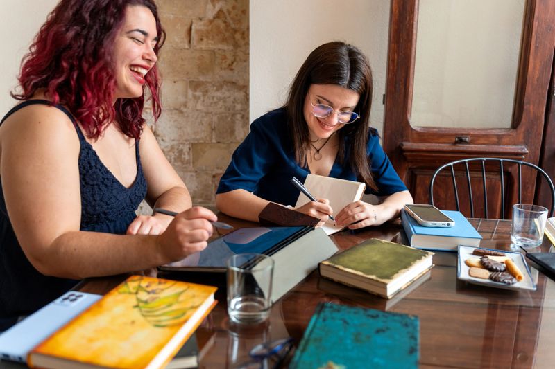 Two female writers smiling and working together on a new book manuscript at home, taking notes and sharing ideas