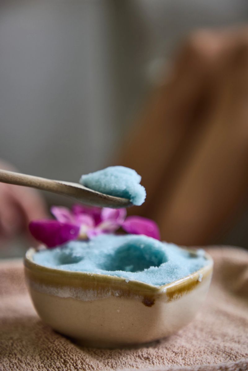 A close-up of a wooden spoon with blue body scrub, a ceramic bowl, and a pink orchid flower nearby, capturing a relaxing and rejuvenating spa or self-care atmosphere with natural elements.