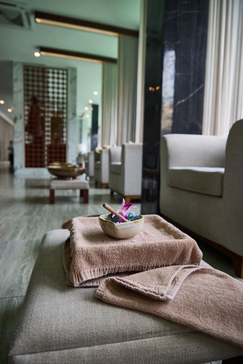 Serene spa setting featuring a neatly arranged towel topped with a bowl containing candles and a flower, against the backdrop of a stylish relaxation lounge with armchairs and soft lighting.