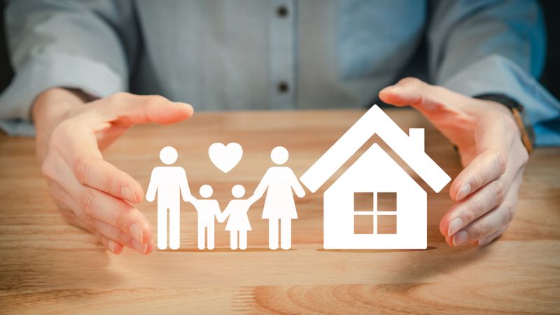 Hands protecting cutout paper family and house icon on wooden table symbolizing home insurance safety and family care concept