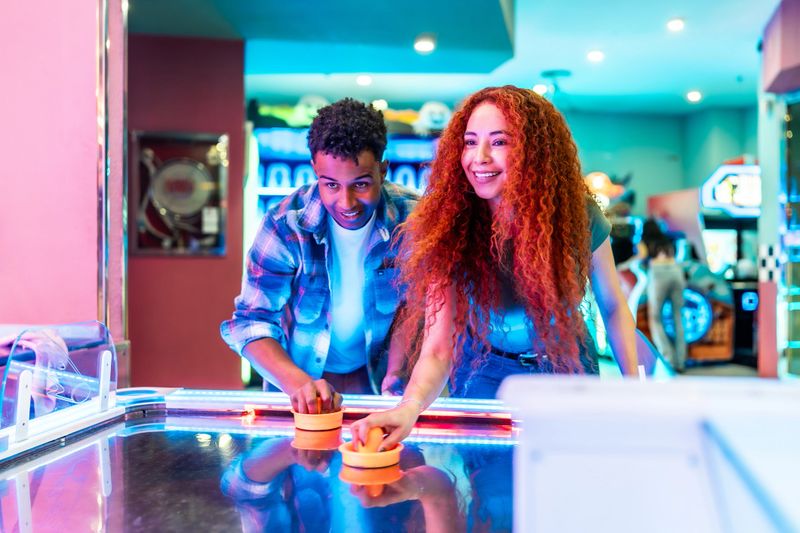 Two young multiethnic friends are enjoying a weekend together, playing air hockey in a brightly lit amusement arcade