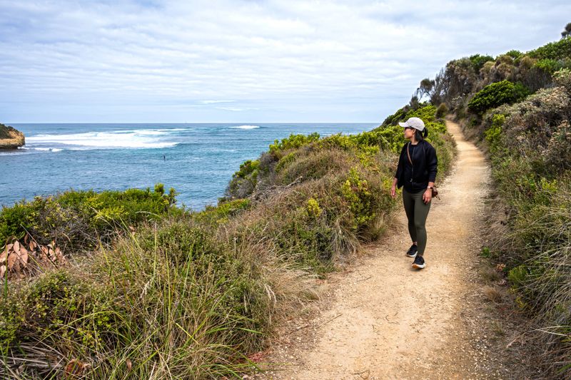 Woman walking on the hiking trail along the ocean, turning her head to see people surfing at the sea, Port Campbell, Great Ocean Road, Victoria, Australia.