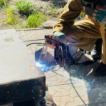Person welding metal outdoors with sparks and smoke visible.