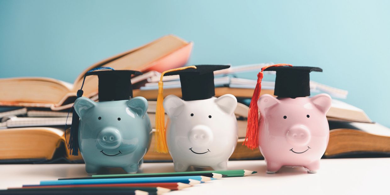 Three piggy banks wearing graduation caps in front of open books and colored pencils.