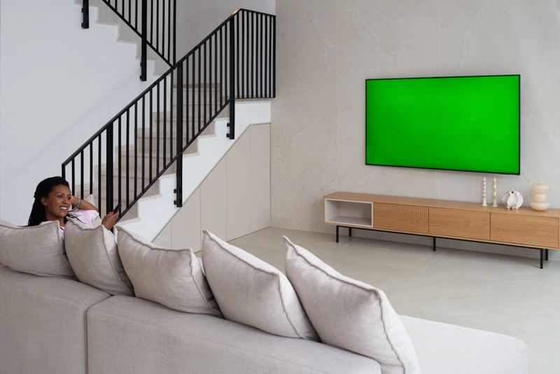 A smiling woman sits on a beige sectional sofa in a stylish, open-concept living room with a staircase and minimalist decor, holding a remote control and facing a large wall-mounted television with a green screen. The scene reflects a blend of contemporary design, smart home comfort, and everyday leisure.