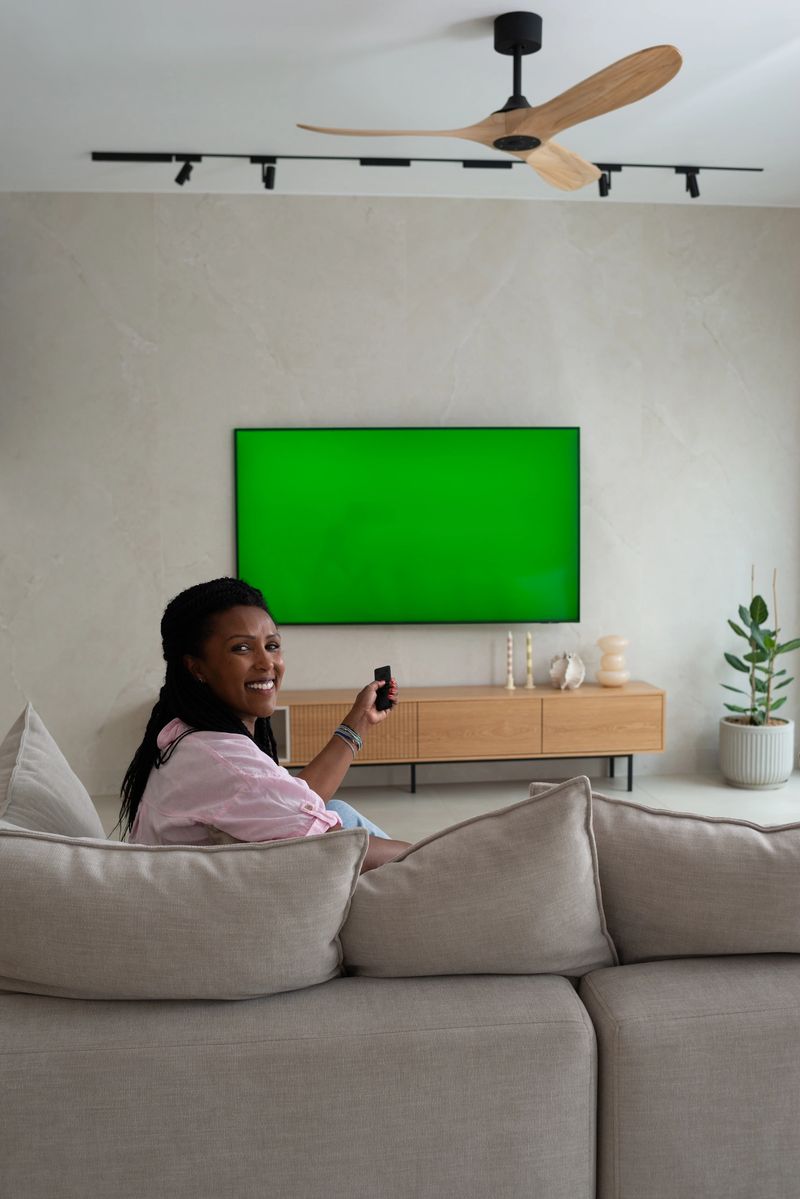 A smiling woman sits comfortably on a beige sectional sofa in a stylish, modern living room. She holds a remote control and faces a large green screen television mounted on the wall. The room features minimalist decor, a wooden media console, an indoor plant, and a wooden ceiling fan, creating a cozy and connected atmosphere.