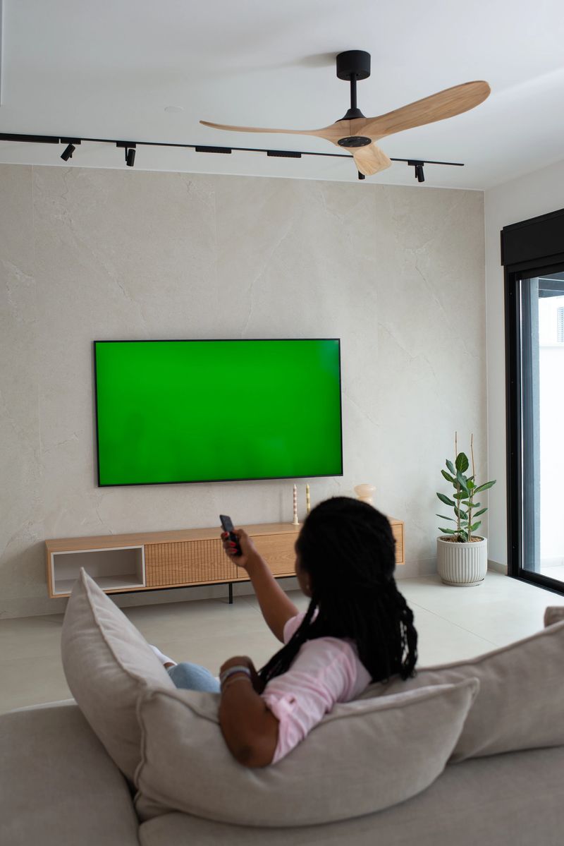 A woman with braided hair is seen from behind, relaxing on a beige sectional sofa while pointing a remote control at a wall-mounted green-screen television. The living room features minimalist decor, a wooden media console, a potted plant, a large window, and a ceiling fan, embodying a serene and tech-savvy home lifestyle.