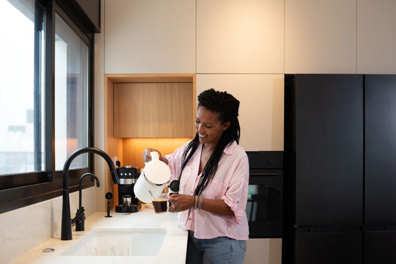 A cheerful woman is seen pouring coffee from a white kettle into a glass cup while standing in a sleek modern kitchen. The natural daylight highlights the minimalist design, black appliances, and light wood cabinetry, creating a warm and contemporary domestic setting.