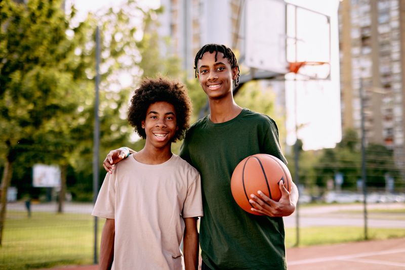 Two friends pose happily on an outdoor basketball court, with one holding a basketball. They portray a moment of friendship and joy, set against an urban cityscape.