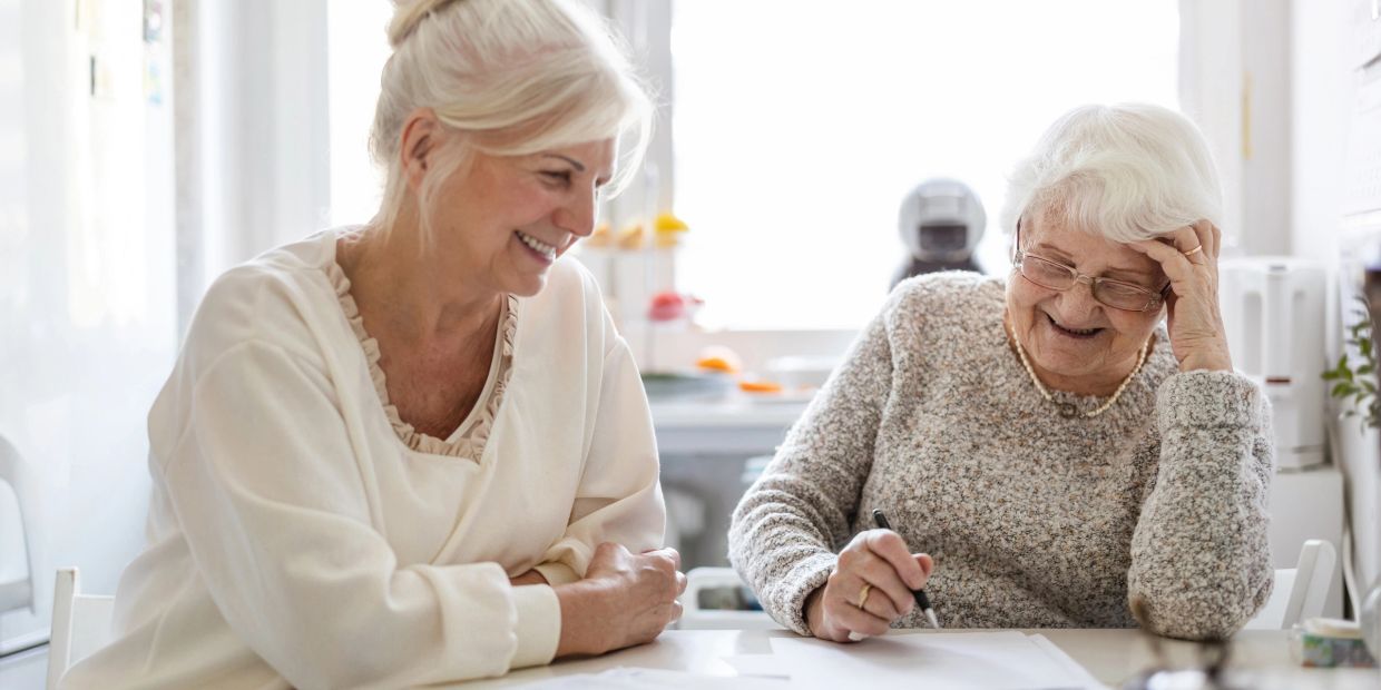 Two elderly women sharing a joyful moment while writing at a table.