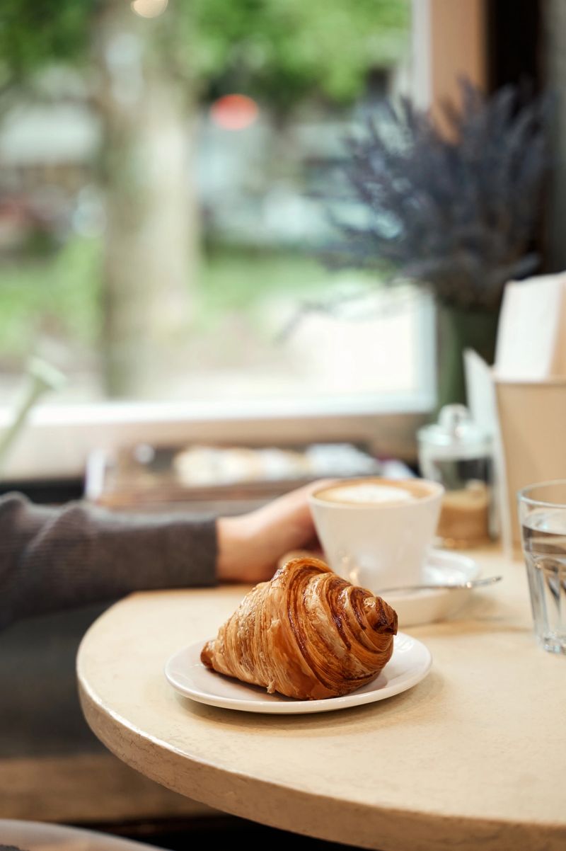 Delicious croissant sits on a plate next to a cappuccino in a cafe, with a customer's hand visible in the background, creating a cozy and inviting atmosphere