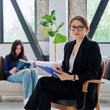 Professional woman holding a clipboard, with a person reading a book in the background.