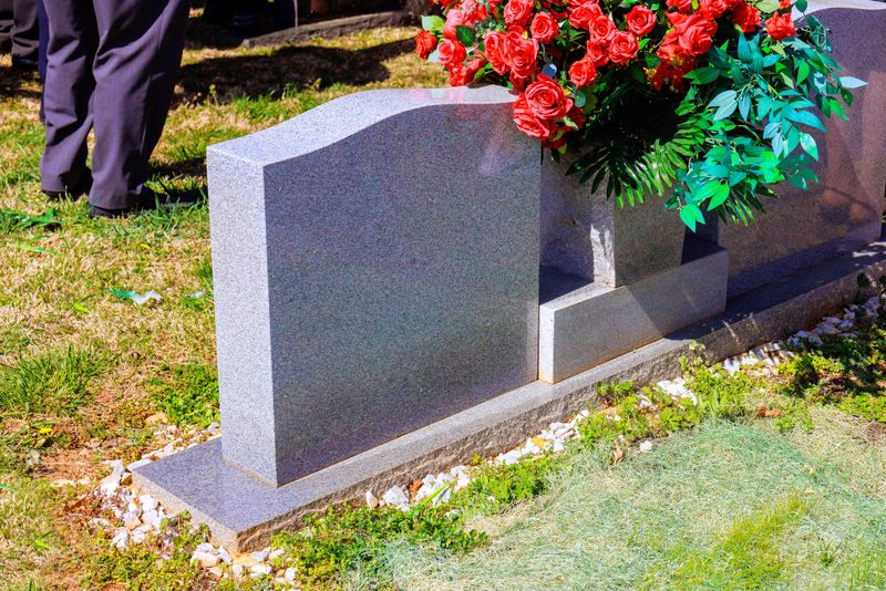 Attendees gather around gravestone adorned with flowers, honoring loved one in peaceful cemetery.