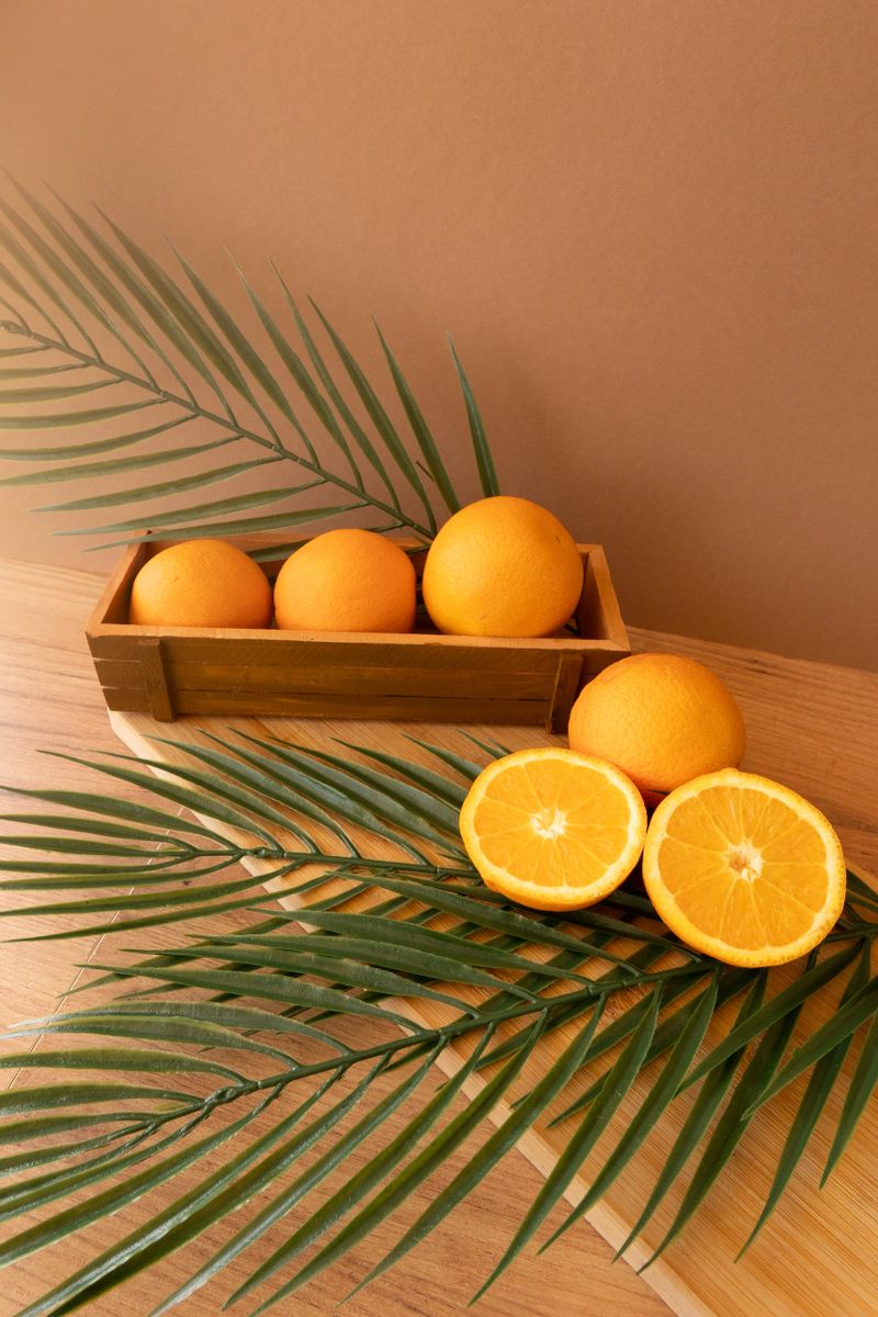 Three whole oranges in a wooden crate and two vibrant halves rest on a wooden board, surrounded by palm leaves for a tropical and wellness-inspired aesthetic filled with freshness, balance and natural