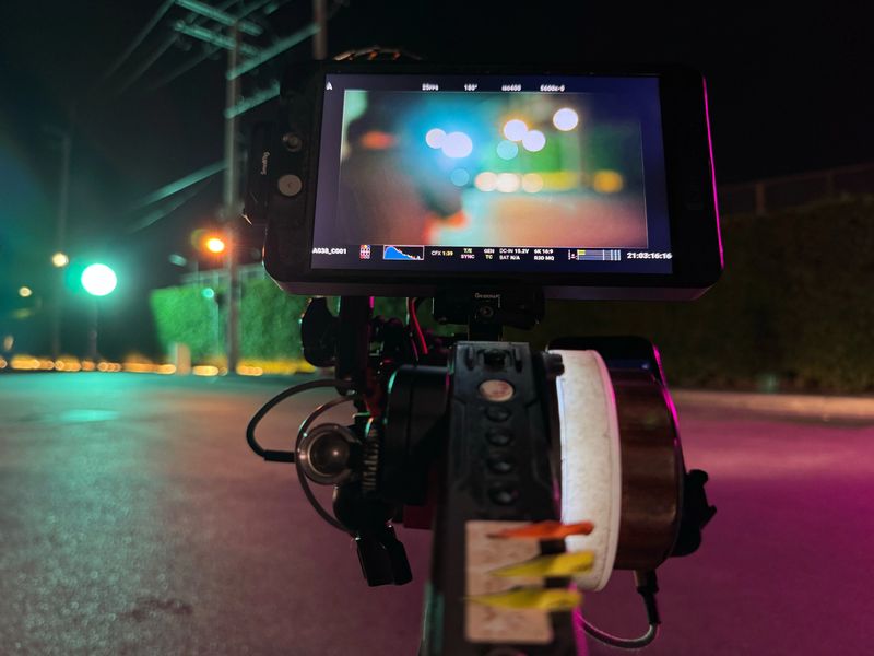 A behind-the-scenes image from a nighttime film shoot, showing a camera monitor mounted on a rig with a follow focus system. The monitor displays a blurred frame filled with colorful bokeh lights, reflecting a cinematic urban street scene. The street is dimly lit, with vibrant hues from streetlights and production lighting casting purples and greens across the pavement. This image captures the technical artistry and atmosphere of filmmaking at night.