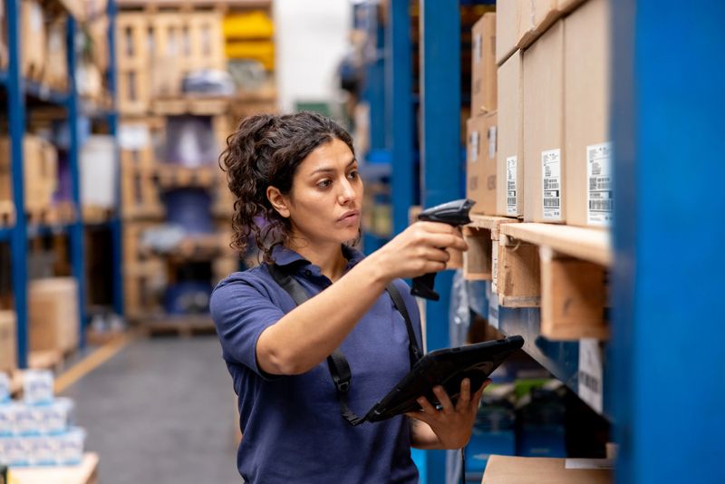 Latin American woman working at a distribution warehouse taking inventory using a bar code reader and a tablet