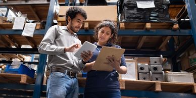 Two workers reviewing inventory on a tablet and clipboard in a warehouse.