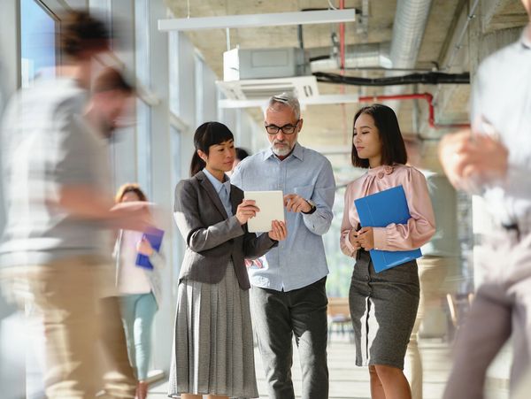 Three professionals discussing work on a tablet in a modern office corridor.