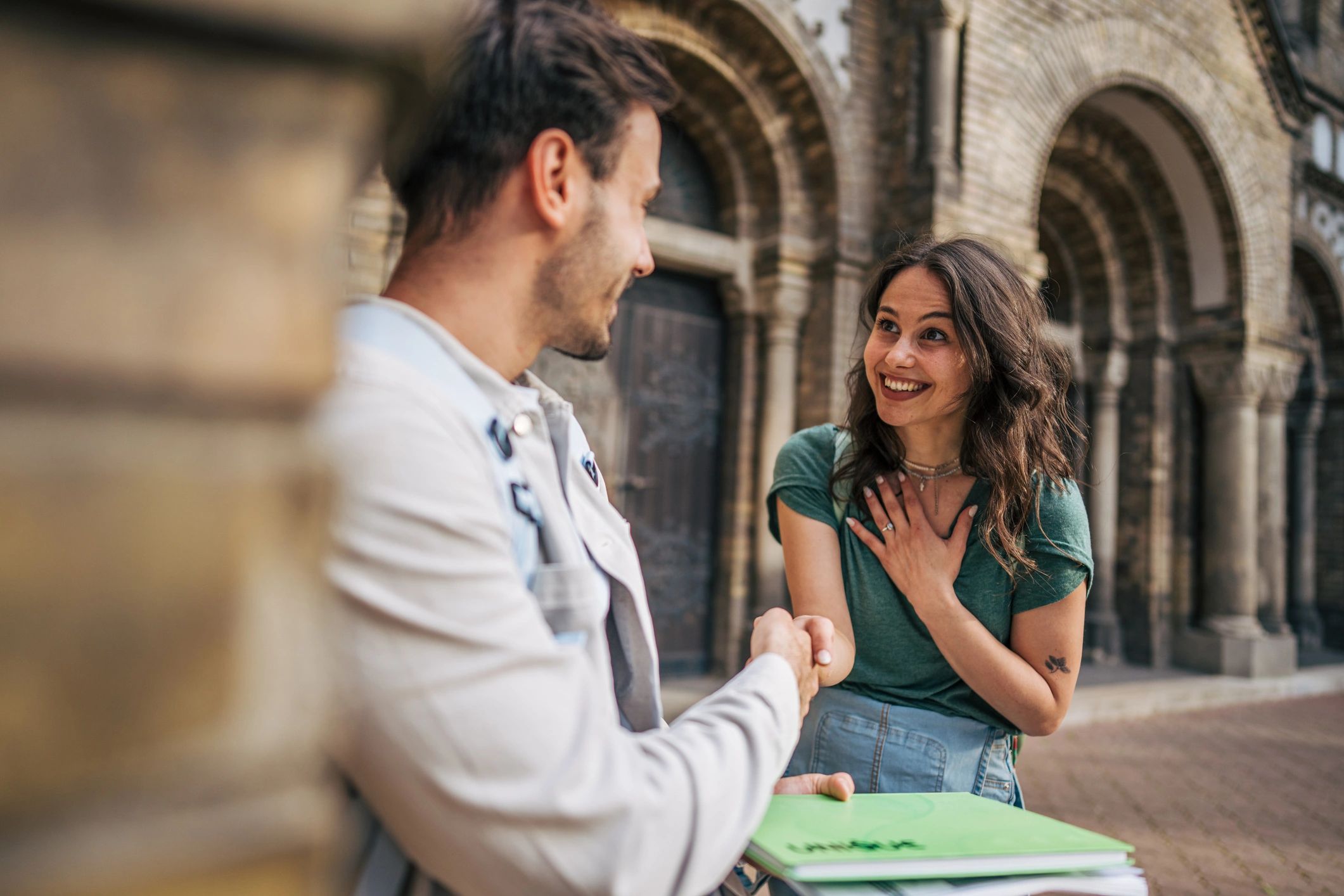 A man and woman smiling and shaking hands outside a building with arches.