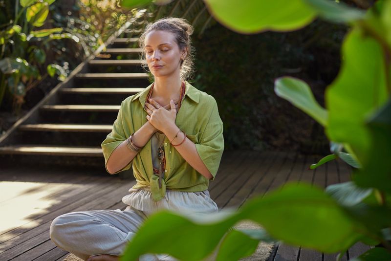 Peaceful woman sits cross-legged in a meditative state, hands over her heart, expressing gratitude and mindfulness in a lush. Girl connects with her emotions during a reflective moment, embracing inner peace. Calm woman practices a heart-centered meditation, cultivating self-love and healing energy, surrounded by greenery.