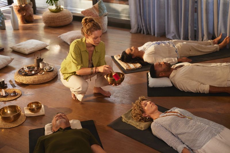 Woman leading sound healing session with singing bowl at indoor wellness retreat. Group of multiethnic people lying down in deep relaxation on yoga mats, surrounded by crystals and warm natural light. Peaceful moment of energy work with people lying on back in circle while the holistic therapist plays tibetan bells.