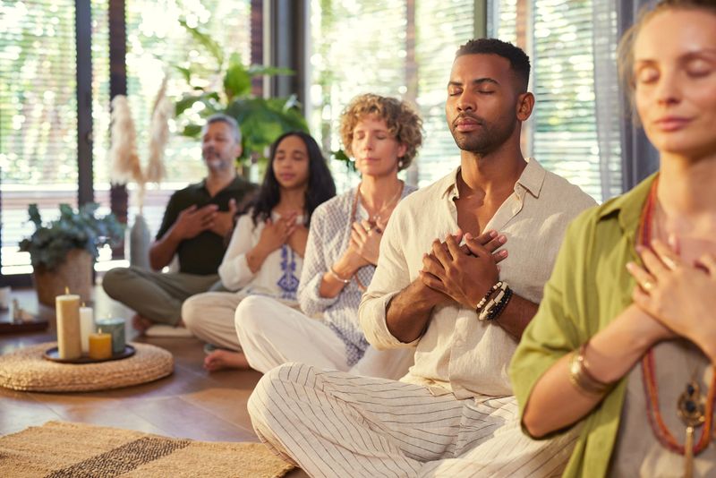 Diverse group meditating with hands on heart at indoor wellness retreat with copy space. Group of people sitting together in a line practicing mindfulness meditation with hand on chest and closed eyes. Peaceful session focused on gratitude, healing, and connection.