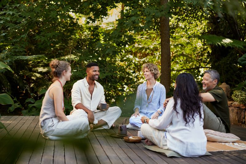 Group of diverse people enjoy each other's company while sitting outdoors at a peaceful wellness retreat. Multiethnic friends relax and talk in a calm natural environment during a detox retreat. Mixed race adults sitting in a circle enjoying tea together in nature.