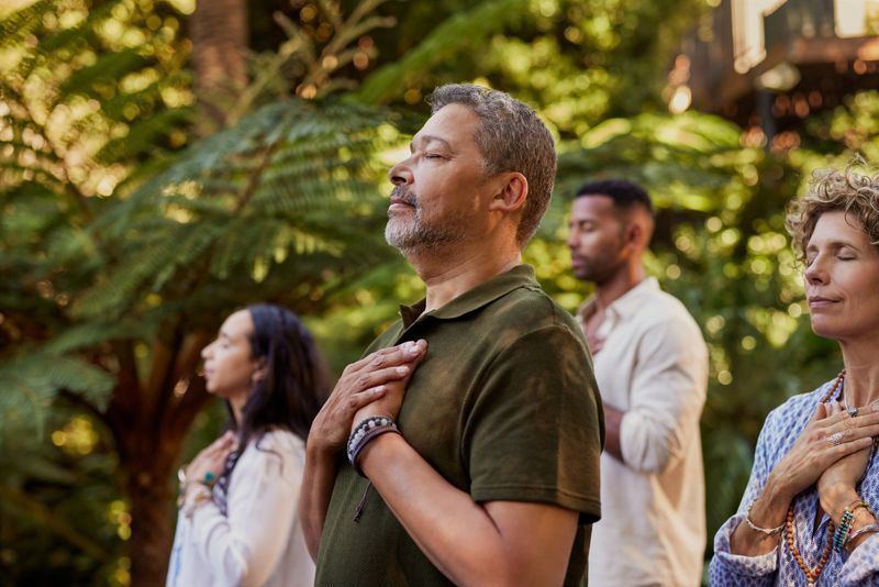 Group of multiethnic people standing outdoors with eyes closed and hands on their hearts during a meditation session. Multiracial adults practicing gratitude and mindfulness together. Mature latin man connecting to inner peace and presence at wellness retreat center in nature.