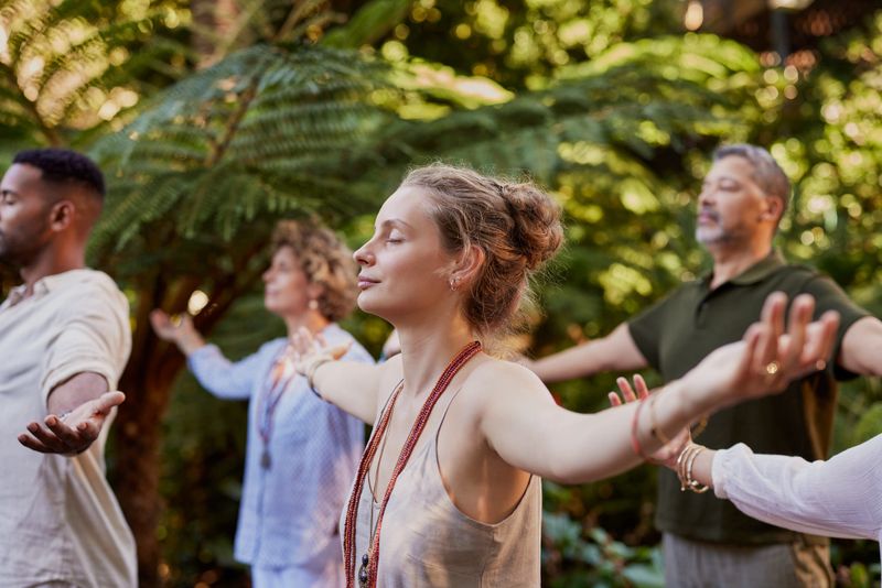 A diverse group practicing mindful breathing with arms outstretched in a peaceful forest setting. Muliethnic people meditating outdoors with eyes closed, focusing on healing and connection. Participants enjoying a wellness retreat surrounded by nature, practicing gratitude and holistic exercises .