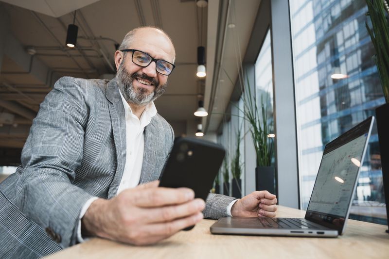 Happy business professional in a modern office, smiling while using smartphone, laptop with stock market charts the desk. The workspace features large windows and natural light