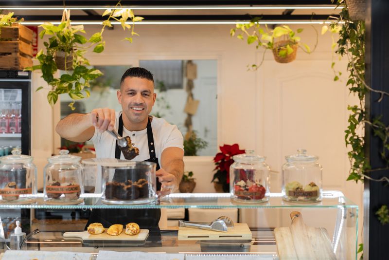 Smiling chef picks a brownie with tongs from a glass jar behind a counter filled with sweet treats. Background is softly blurred. Horizontal medium shot, copy-space