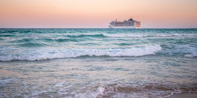 Cruise ship sailing at sunset off a sandy beach with gentle waves.