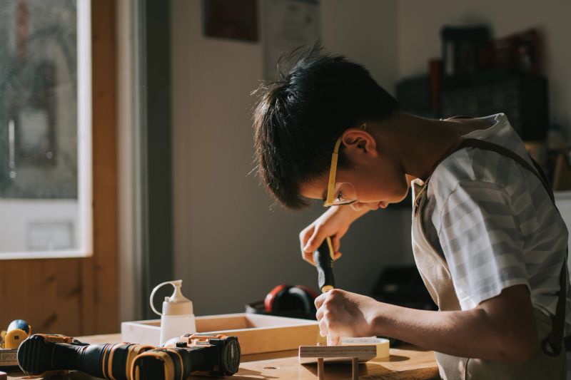 young asian chinese boy wearing safety glasses and an apron concentrates on a woodworking project at a sunlit indoor workshop. Surrounded by tools and natural light, the scene highlights creativity, focus, and hands-on learning. Perfect for themes around education, DIY, craftsmanship, or childhood development.