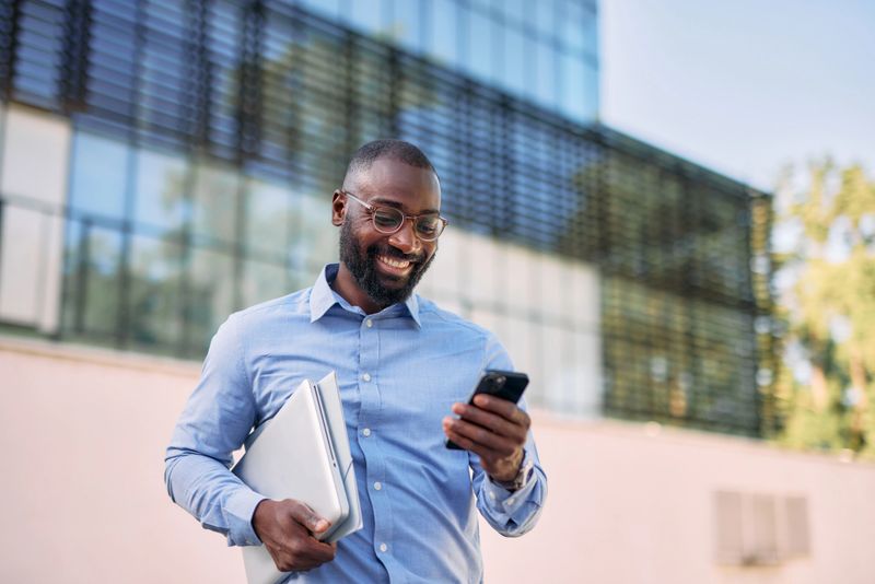 Cheerful African American businessman walking confidently in front of an office building. Carrying a laptop while engaging in a conversation on his smartphone. Exuding professionalism and positivity