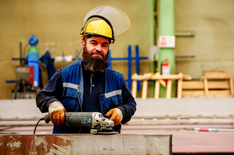A skilled industrial worker wearing protective gear performs  using a grinder on a large metal pipeline in a fabrication workshop.
