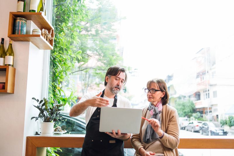 Cafe owner consults with financial adviser using laptop in a bright cafe setting. They collaborate on strategy, showcasing small business growth, digital planning, and entrepreneurial teamwork.