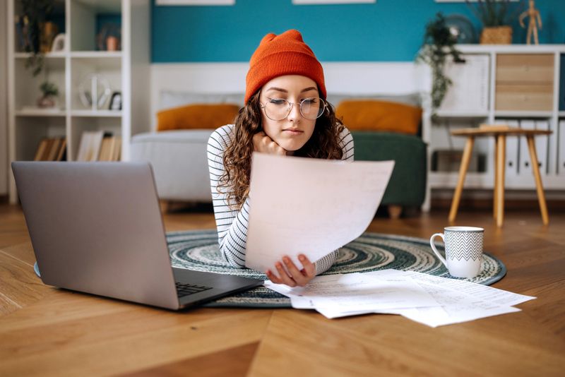 A young woman lies on her stomach on the floor of her cozy apartment, deeply focused on her studies as she uses her laptop