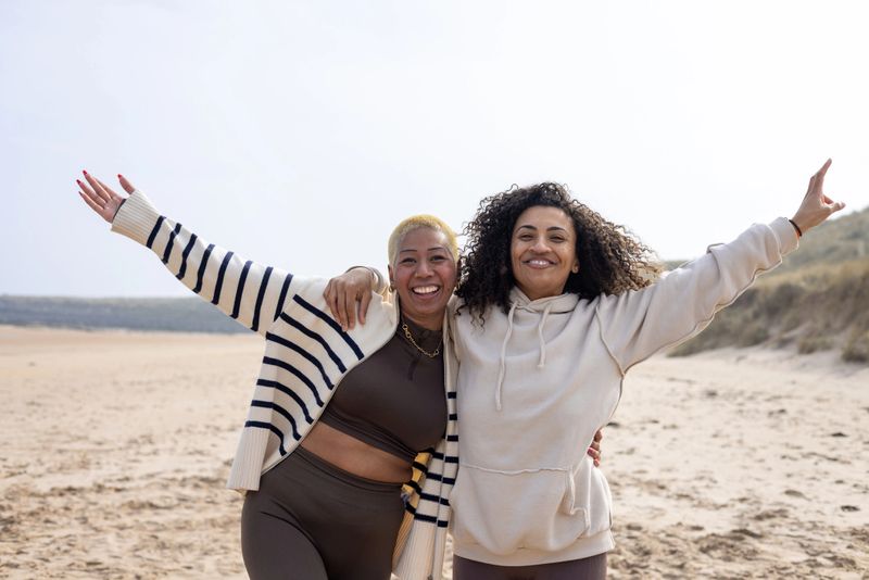 Medium shot of two female friends embracing on the beach in Northumberland, North East England. They are both smiling directly at the camera with their outer arms outstretched. They are wearing casual clothing and sand dunes are visible in the background.

Videos similar to this scenario available.