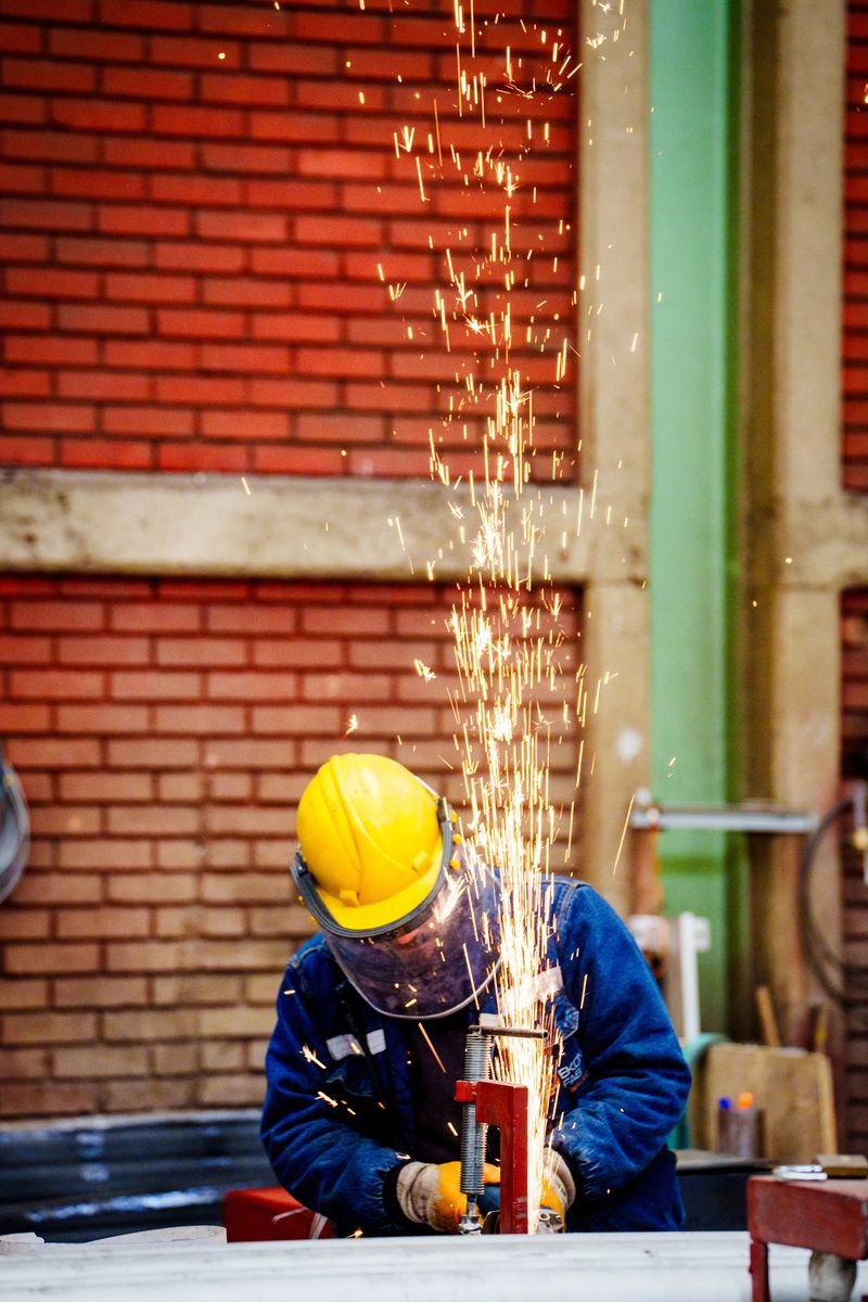 A skilled industrial worker wearing protective gear performs TIG (Tungsten Inert Gas) welding on a large metal pipeline in a fabrication workshop. The welder uses precision techniques while the pipe is secured with a clamp. Bright welding sparks and industrial tools are visible in the background, highlighting the heavy manufacturing environment. Concept of skilled labor, industrial engineering, metalwork, and mechanical fabrication.