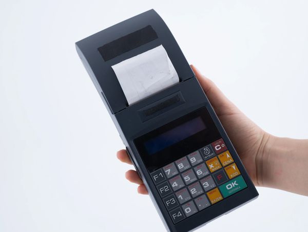 A person's hand holding a dark grey, handheld terminal/printer that is dispensing a small receipt.