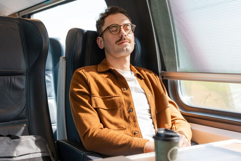 A man wearing glasses and a brown jacket relaxes during a train journey. He has a calm expression, seated by the window. Captures the concept of relaxation during travel.