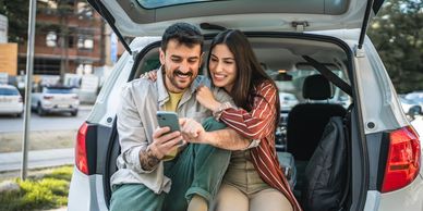Couple sitting in a car trunk, happily looking at a phone together.