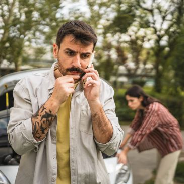 A man talks on the phone looking worried while a woman sits distressed in the background.