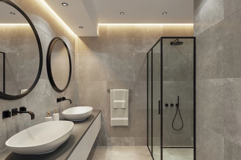 Spacious and elegant modern bathroom featuring a long floating counter top with two white oval vessel sinks and matching round mirrors. Walls and floor are covered with large-format natural stone-effect tiles in soft grey tones, creating a calming, spa-like atmosphere. A glass shower enclosure with a black metal frame adds contrast, while built-in LED spotlights and a concealed LED strip above the sinks provide layered lighting. Ideal for promoting ceramic tile manufacturers, bathroom renovation projects, interior design studios, architecture firms, and contemporary wellness spaces.