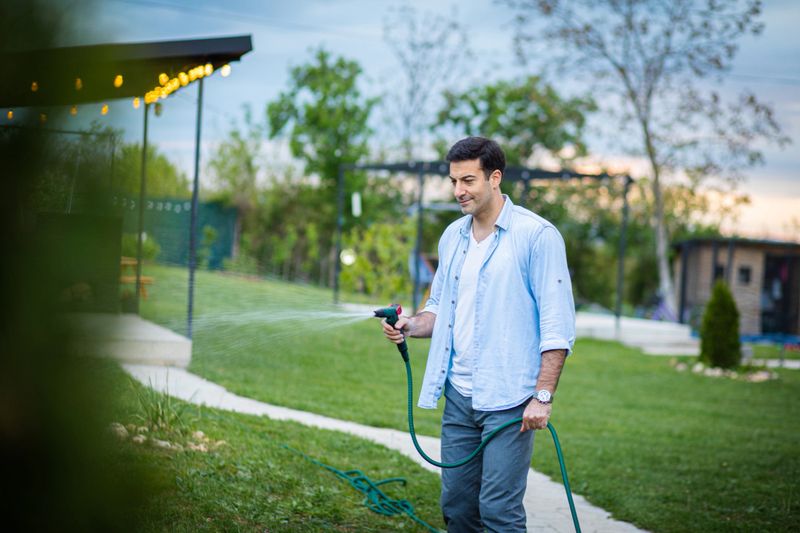 Handsome man watering green lawn, sprinkling water on the grass in backyard