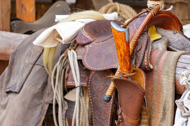 detailed close-up of a vintage Western cowboy saddle with intricate leather tooling, a tomahawk in a leather holster, coiled ropes, and rustic fabrics draped over a wooden structure. Captures the spirit of the Wild West—perfect for use in historical, western-themed, or rustic lifestyle projects.
