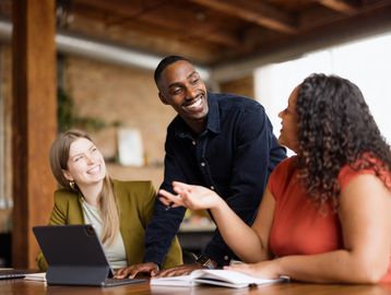 Three colleagues smiling and discussing work around a table with a tablet and notebook.