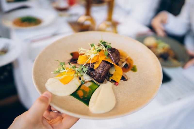 A hand presents a beautifully arranged gourmet dish featuring tender meat with vibrant vegetables and creamy sauce at a fine dining restaurant during dinner time.